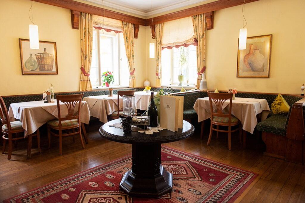 Cozy restaurant interior with wooden floors and warm lighting in the sustainable Hotel Schloss Lerchenhof in Gailtal, Austria. Round table with wine bottles and menus in the center. Windows with curtains, surrounded by tables with white tablecloths. Warm, inviting atmosphere.