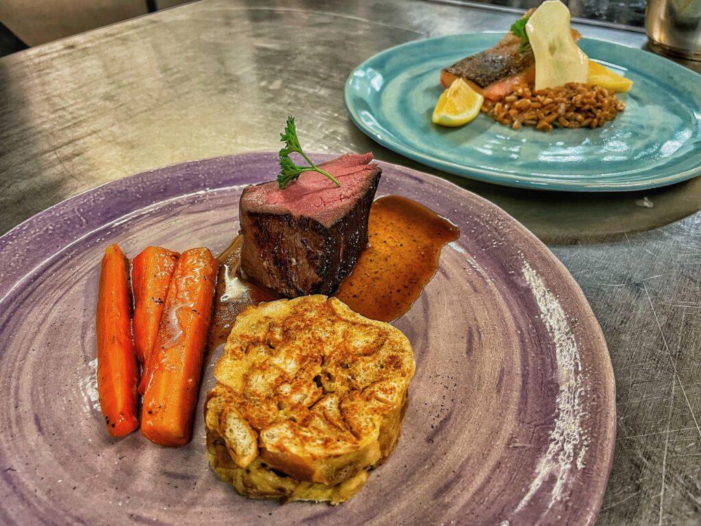 Regional gourmet dishes on a table in the Slow Food restaurant at the Hotel Schloss Lerchenhof in Carinthia: in the foreground, a plate of steak, carrots, and potatoes on a purple plate; in the background, salmon with rice on a blue plate.