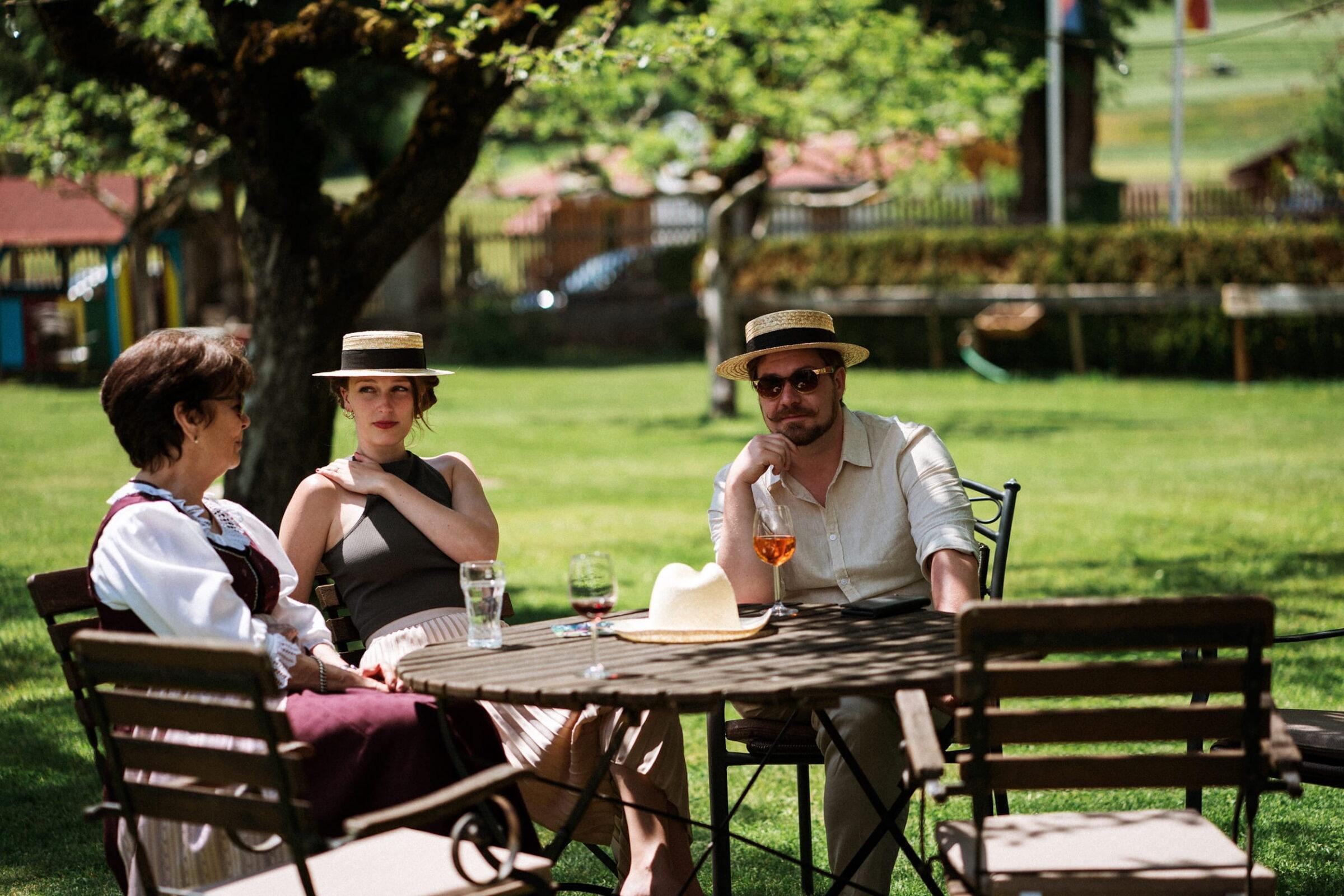 Three people are sitting at a table outside in the sunny garden of the sustainable Hotel Schloss Lerchenhof in Gailtal, Austria, wearing straw hats and sipping their drinks. The atmosphere is relaxed and leisurely.
