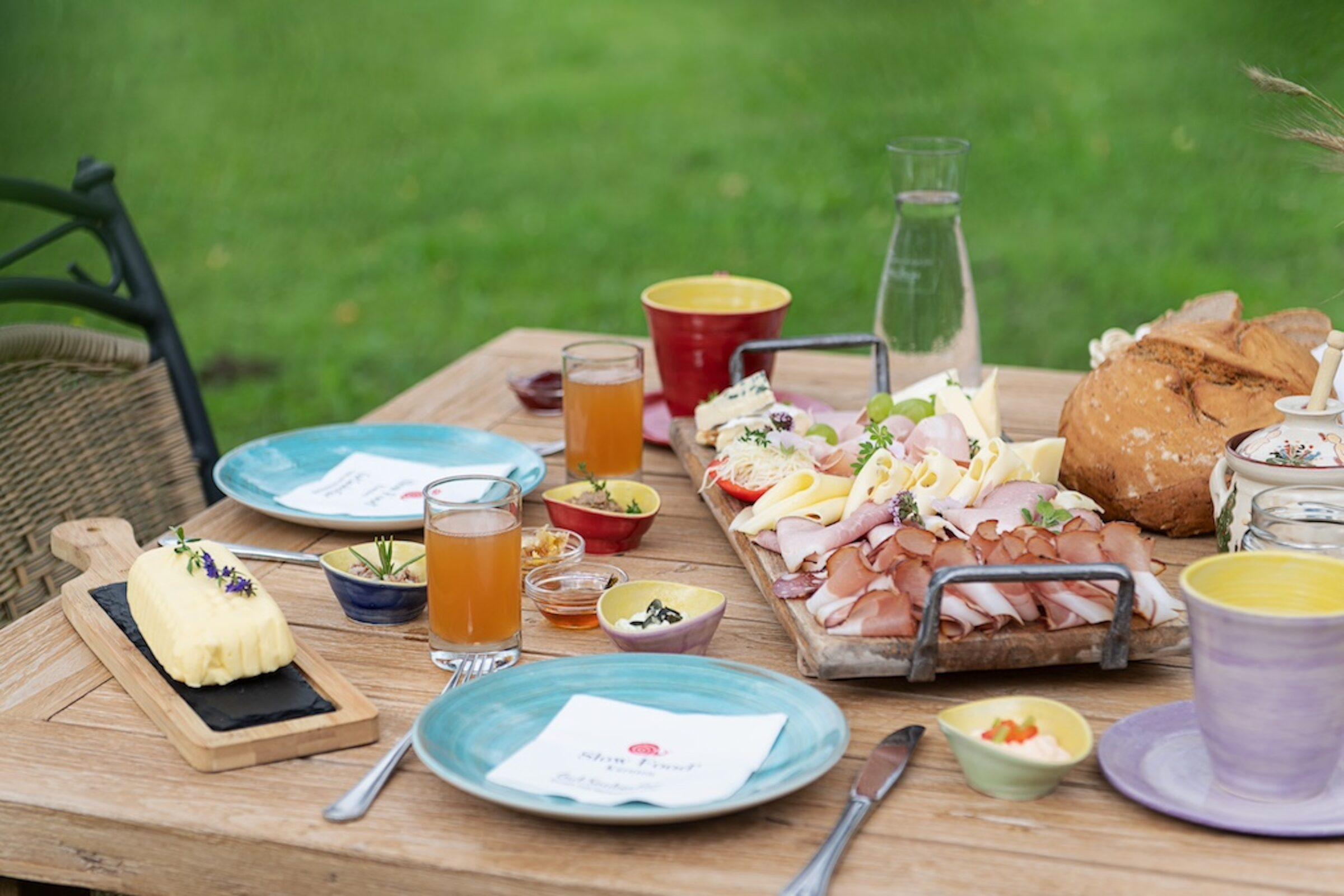 Slow Food picnic on a wooden table in the garden of the sustainable Hotel Schloss Lerchenhof in Austria with colorful plates, ham and cheese platter, rustic bread, herb butter, small bowls, and juice. Green lawn in the background.