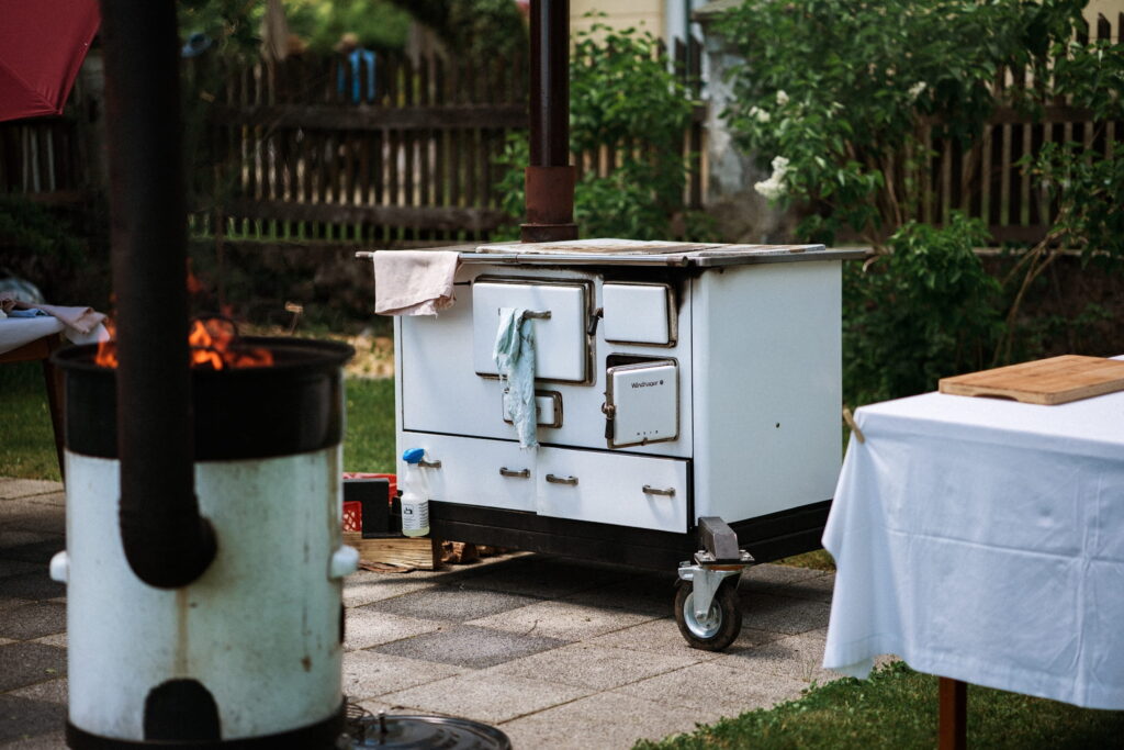 A white vintage stove on wheels outdoors, with a towel draped over its handle. A fire burns nearby under a chimney. The scene has a relaxed, rustic feel. Taken at the sustainable Hotel Schloss Lerchenhof in Carinthia.