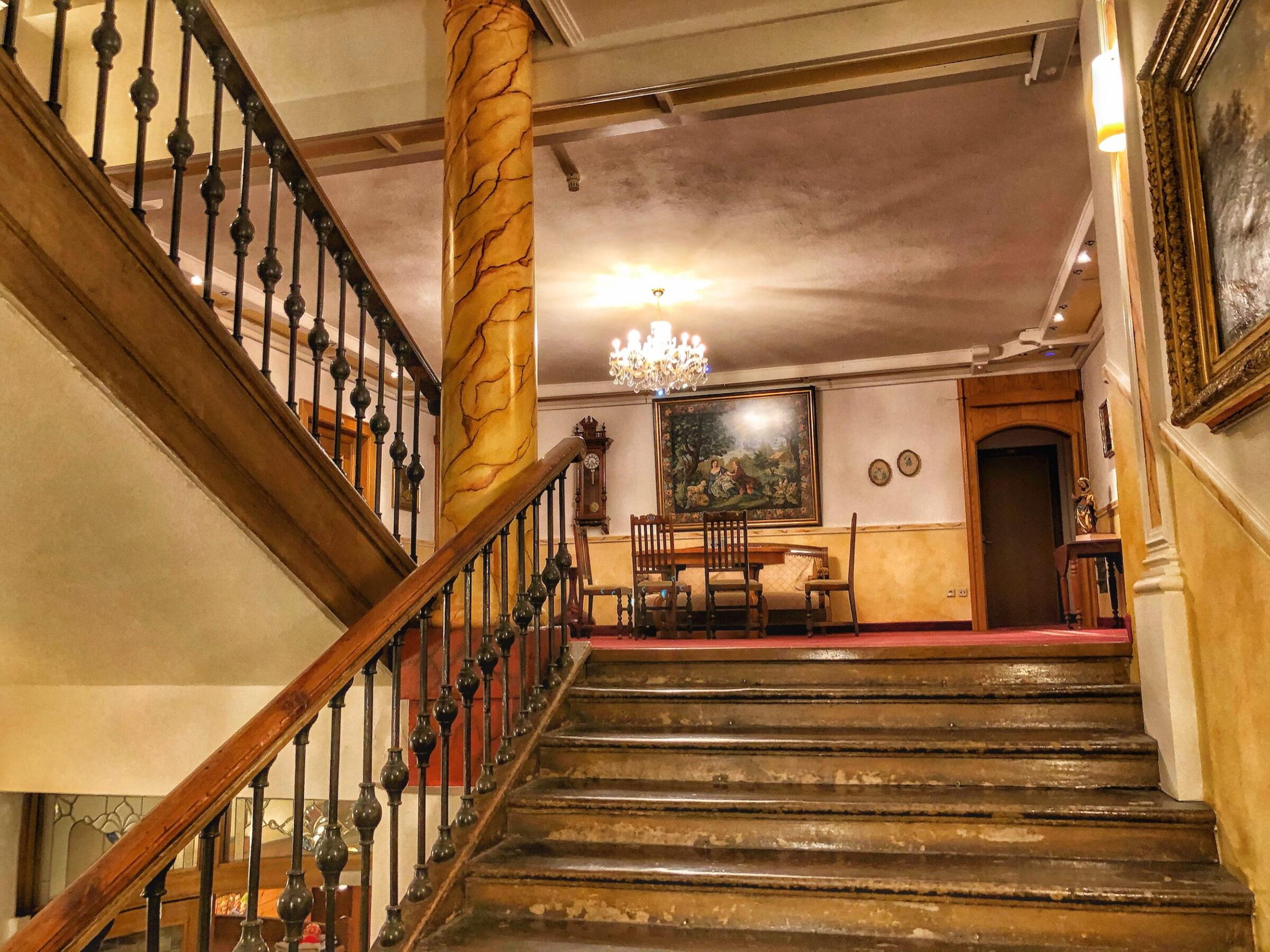 Elegant interior of the sustainable Hotel Schloss Lerchenhof with a large, historic staircase leading to a vintage hall. Warm lighting, a chandelier, and classic artwork create a cozy, nostalgic atmosphere.