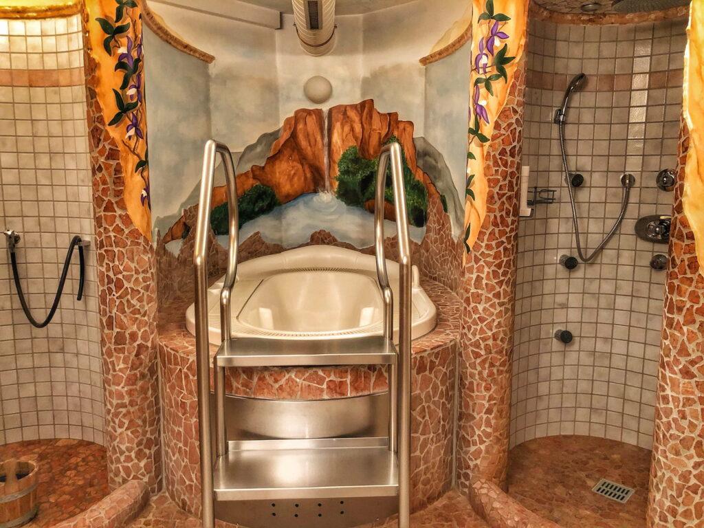 Luxurious wellness room in the sustainable Hotel Schloss Lerchenhof with whirlpool, surrounded by a stone tile design. A mural depicting a waterfall and mountains enhances the calming atmosphere.