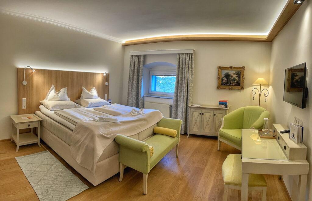 A cozy hotel room in the sustainable boutique hotel Schloss Lerchenhof with a king-size bed, illuminated headboard, and wooden floor. There is a small sofa, an armchair, a desk, wall decorations, and soft lighting.
