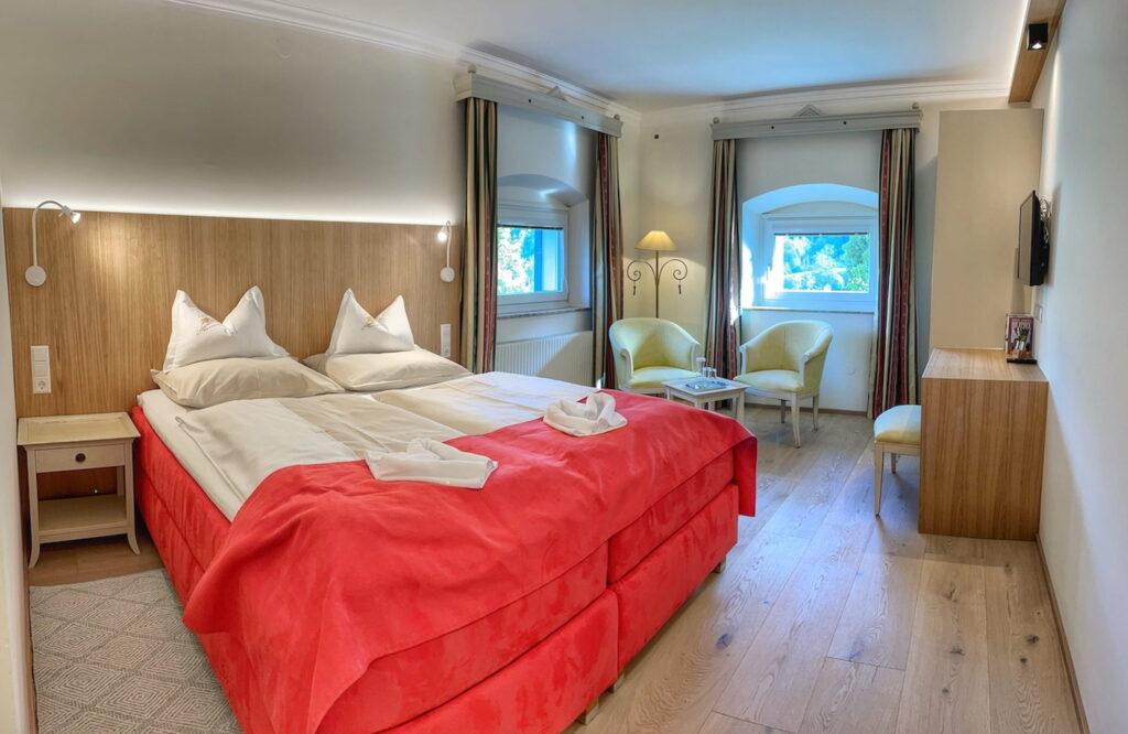 Cozy hotel room in the sustainable Hotel Schloss Lerchenhof in Carinthia with a double bed with red bedding and white pillows. Two yellow chairs and a small table stand next to arched windows, creating a bright, inviting space.