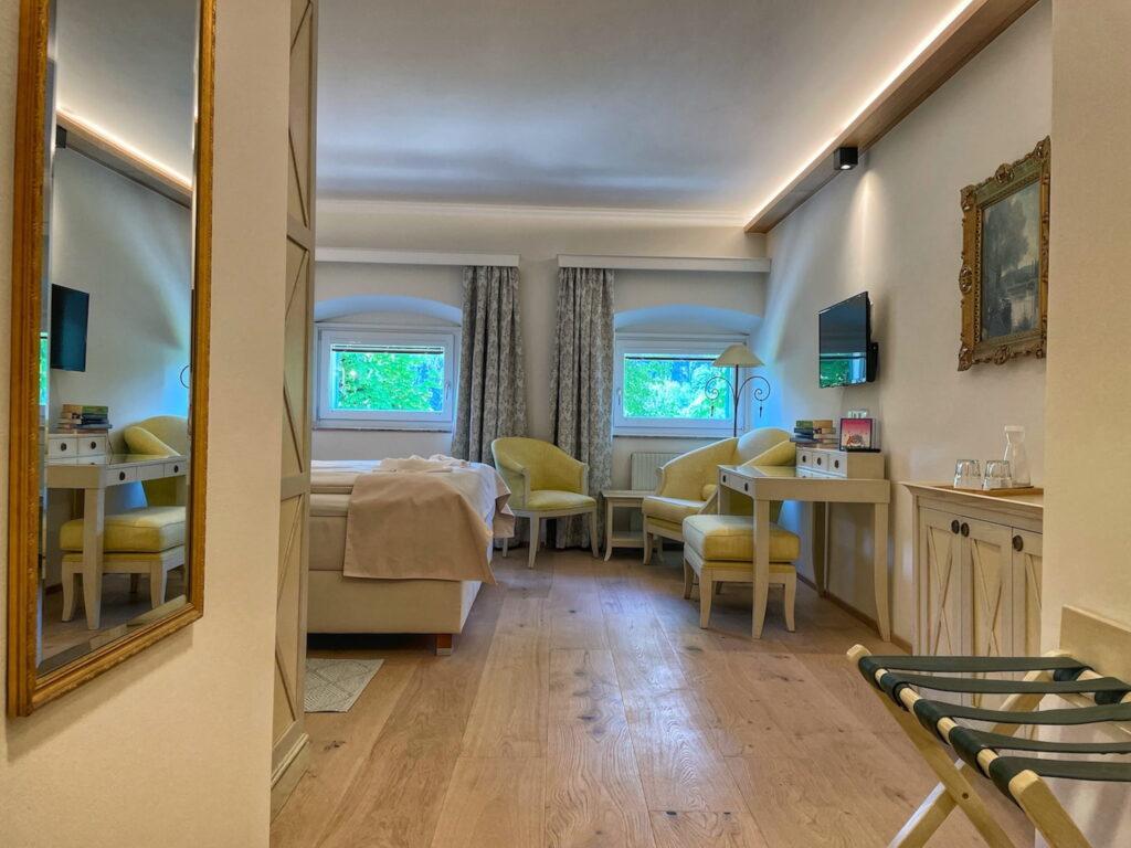 A cozy hotel room in the sustainable Schloss Lerchenhof in Carinthia, Austria, with light wood flooring, soft lighting, and two small windows decorated with floral curtains. It features a neatly made bed, yellow chairs, a wooden desk, a mirror, and a wall-mounted TV, creating a welcoming and relaxed atmosphere.