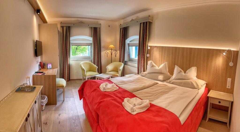 A cozy hotel room at the sustainable Austrian Hotel Schloss Lerchenhof features a neatly made double bed with red blankets and white pillows. Two windows flanked by striped curtains allow soft light to flood into the room, accentuating the light wood flooring. In the corner, two light green armchairs and a lamp create an inviting seating area. A small desk with a wall-mounted TV completes the warm, inviting atmosphere.