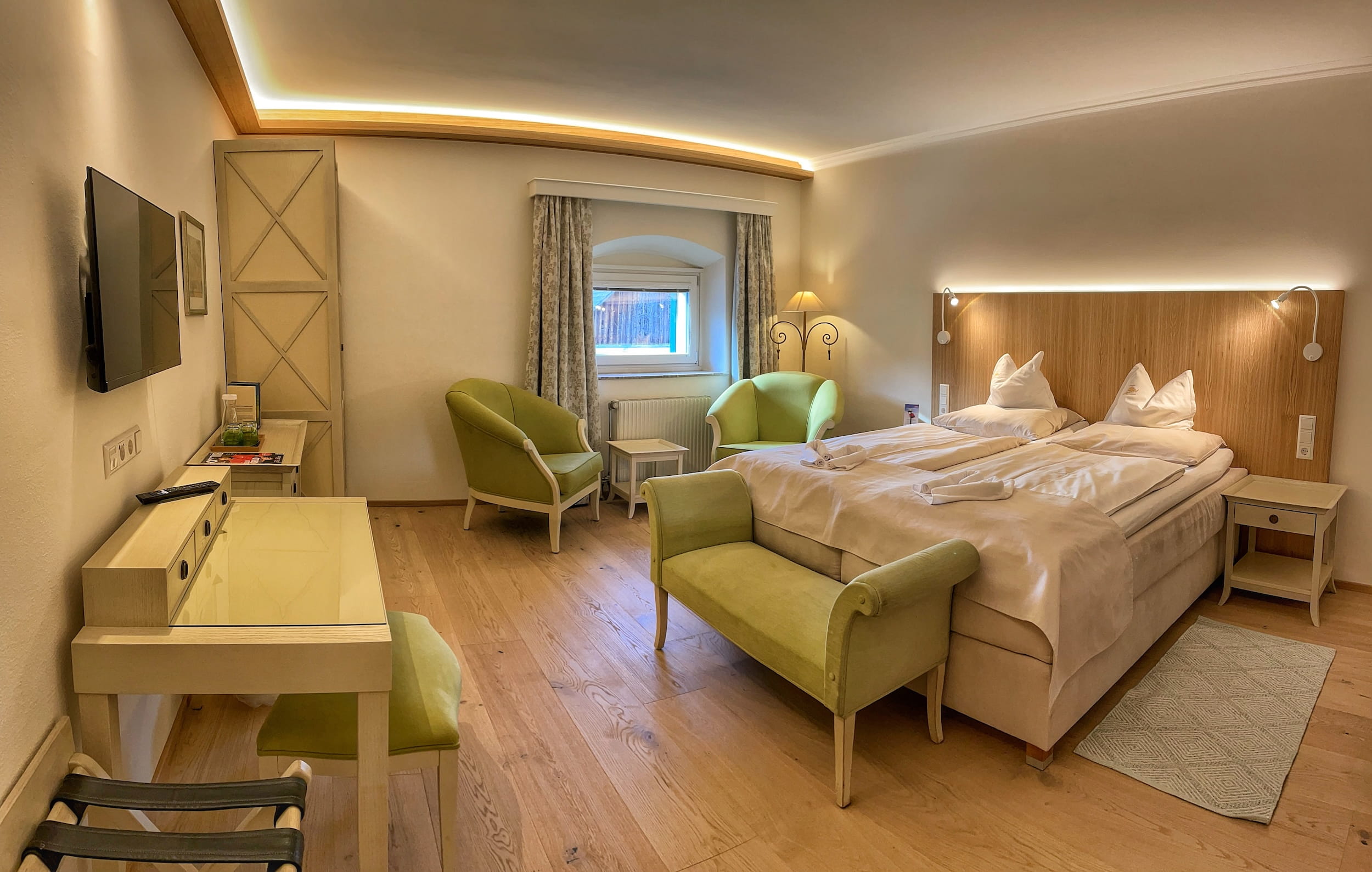 Cozy hotel room at Schloss Lerchenhof in Carinthia, Austria, with a modern, warm atmosphere. Features a large bed with beige bedding, matching armchairs, a small desk, and soft lighting.