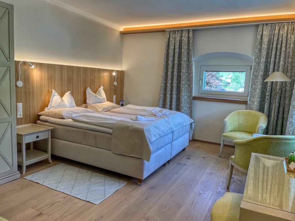 Cozy hotel room in the sustainable boutique hotel Schloss Lerchenhof in Carinthia with a soft double bed, wooden headboard, soft lighting, and elegant furnishings. A small window with a view of the countryside creates a peaceful atmosphere.