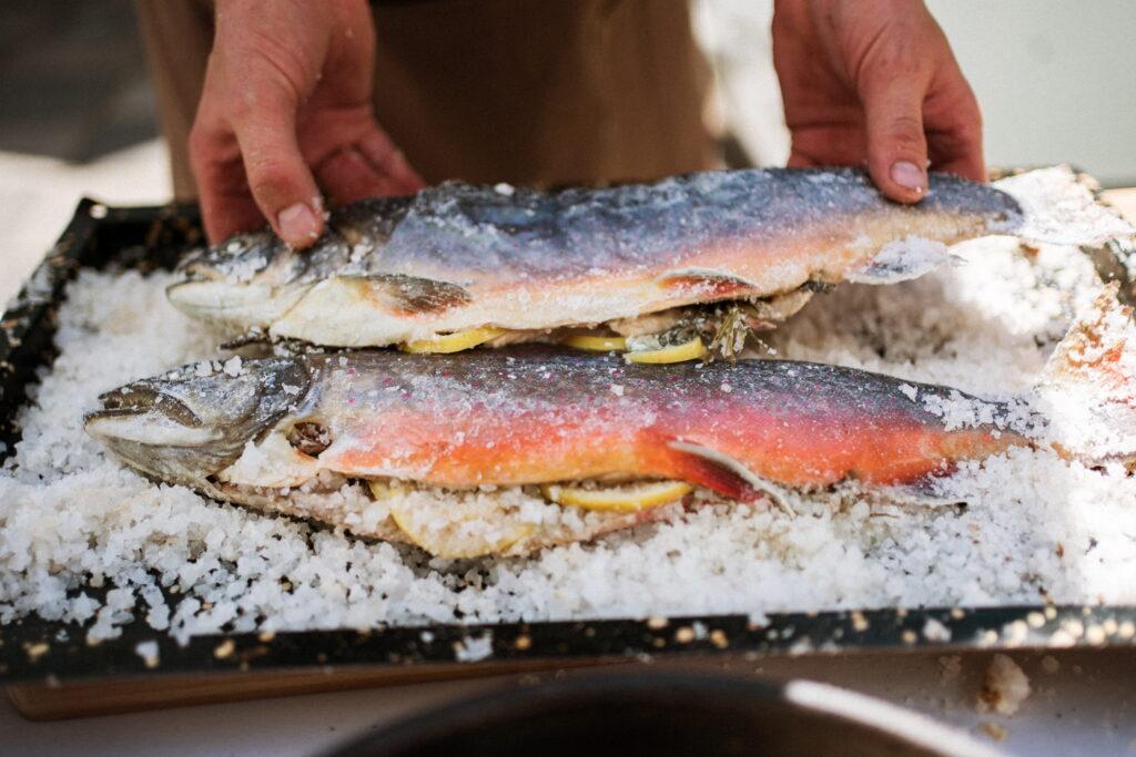 Two fresh fish with bright orange hues are prepared with lemon slices and herbs and placed on a bed of salt. The fish is handled with care, conveying the attention to detail and freshness that is typical of the Austrian Slow Food Hotel Lerchenhof.