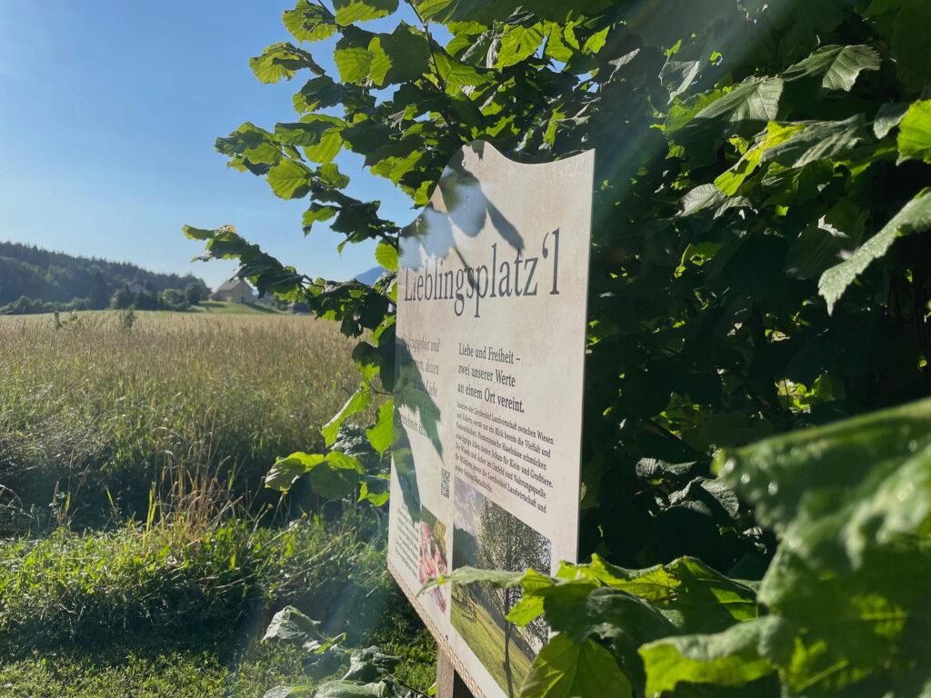 A sunlit sign reading “Lieblingsplatz'l” (favorite spot) in the nature park surrounding the sustainable Hotel Schloss Lerchenhof is partially obscured by lush green leaves. Behind it lies a tranquil landscape of fields and distant hills under a bright blue sky.