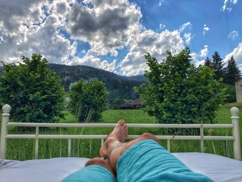 A person is lying on a bed outdoors wearing turquoise shorts. Bright green trees, lush grass, and a mountain under a cloudy blue sky create a peaceful atmosphere at the power spot of the Hotel Schloss Lerchenhof in Austria.