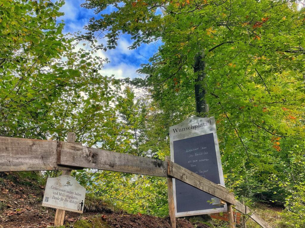 A wooden fence and a sign stand amid lush green trees under a partly cloudy sky. The scene conveys a peaceful and inviting atmosphere at the sustainable Hotel Schloss Lerchenhof in Austria.