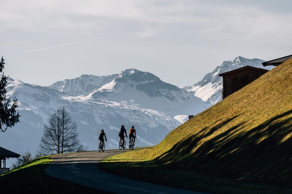 Drei Radfahrer fahren auf einem Weg mit schneebedeckten Bergen der Kitzbüheler Alpen im Hintergrund und einer grasbewachsenen Hanglage rechts