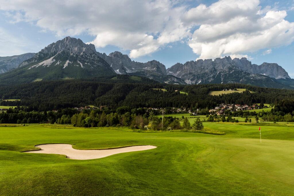 Golfplatz mit Sandbunker und Fahne vor bewaldeten Hügeln und markanten Berggipfeln des Wilden Kaisers in Tirol