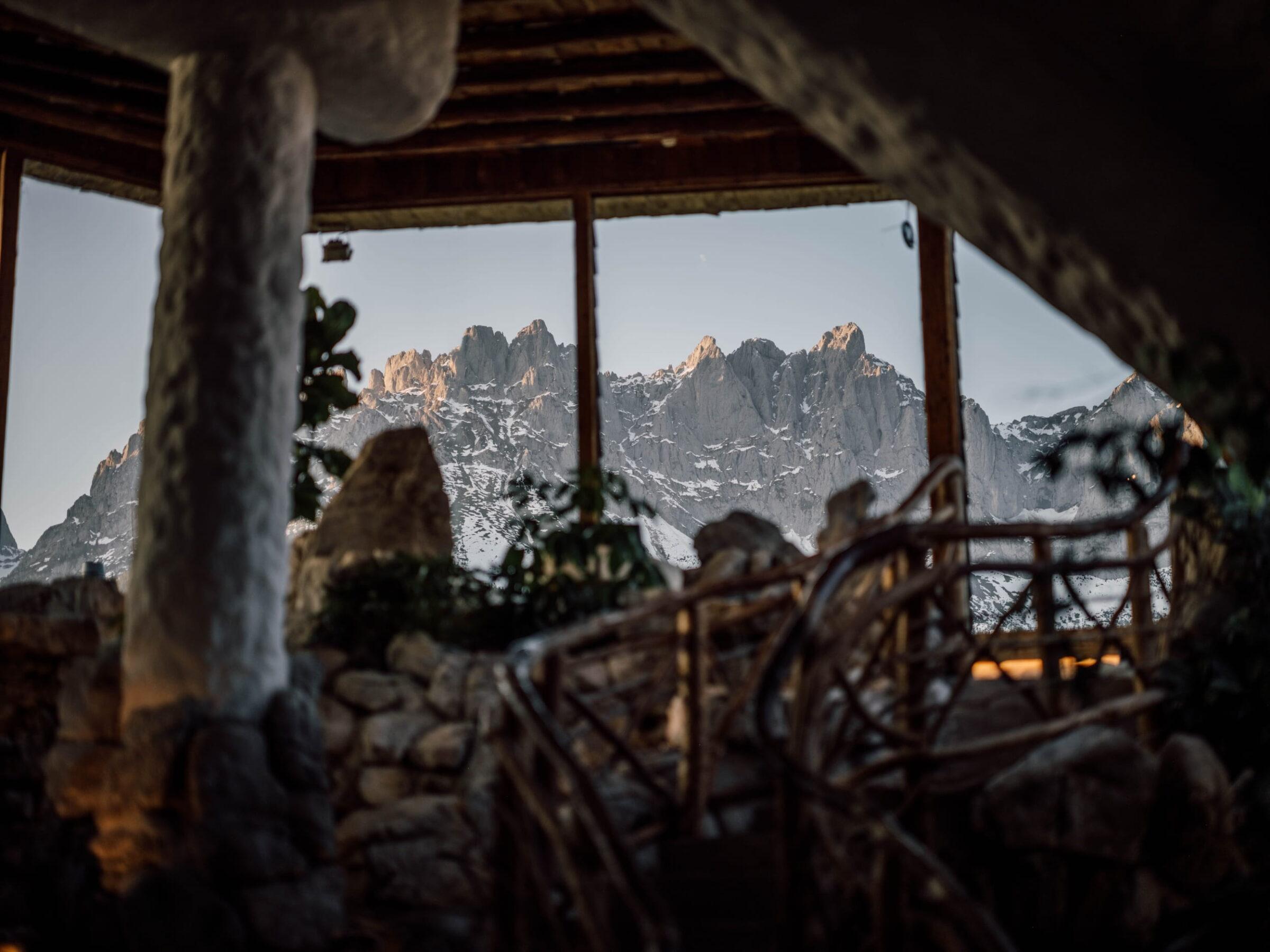 Blick aus einem rustikalen Innenraum im Wellnessresort Stanglwirt mit Naturstein und Holz auf die schroffen Gipfel des Wilden Kaisers bei Abendlicht