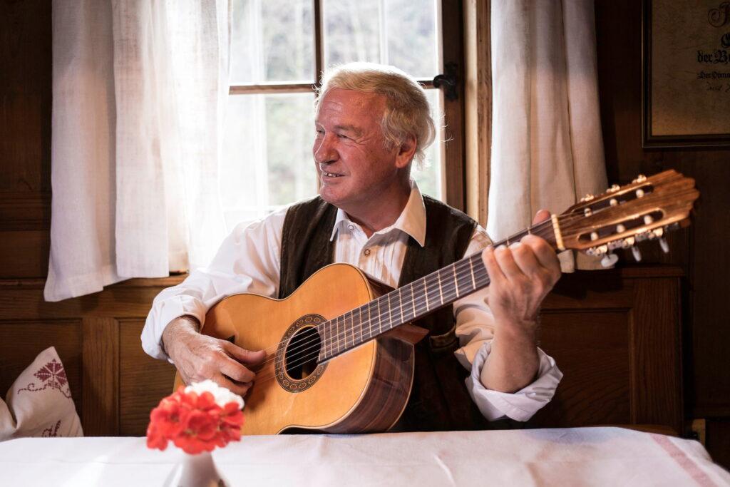 Balthasar Hauser, Inhaber des Stanglwirts, mit weißem Hemd und dunkler Weste sitzt an einem Holztisch und spielt akustische Gitarre vor einem Fenster mit weißen Vorhängen in einem rustikalen Raum