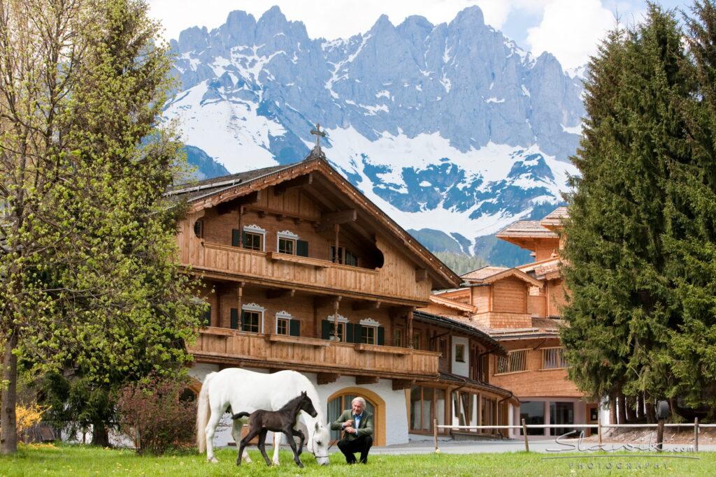 Mann kniet neben weißem Pferd und Fohlen vor dem Holzhaus des Stanglwirts in Tirol mit Blick auf schneebedeckten Berg im Hintergrund