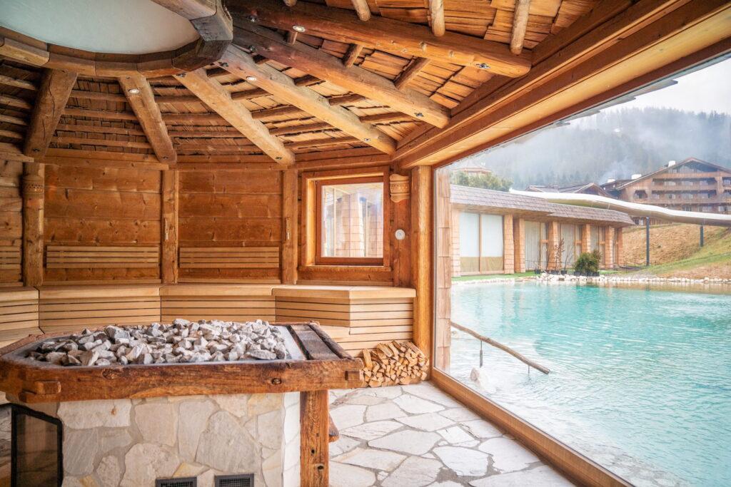 Innenraum einer Sauna aus Holz mit Steinofen und Blick auf einen Naturbadesee und umliegende Gebäude im Bio- und Wellnessresort Stanglwirt in Tirol