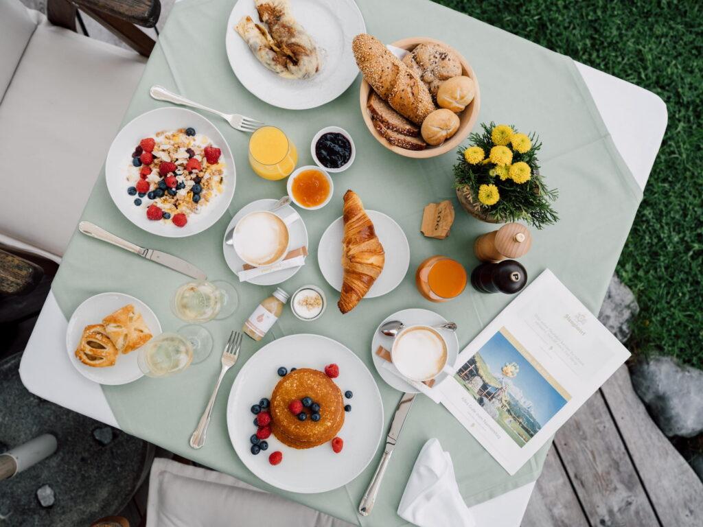 Frühstückstisch im Bio- und Wellnessresort Stanglwirt mit Croissant, Brötchen, Pancakes mit Beeren, Müsli mit Beeren, Kaffee, Orangensaft, Marmelade, Gebäck und einem kleinen Blumenarrangement auf grünem Tischdecke im Freien