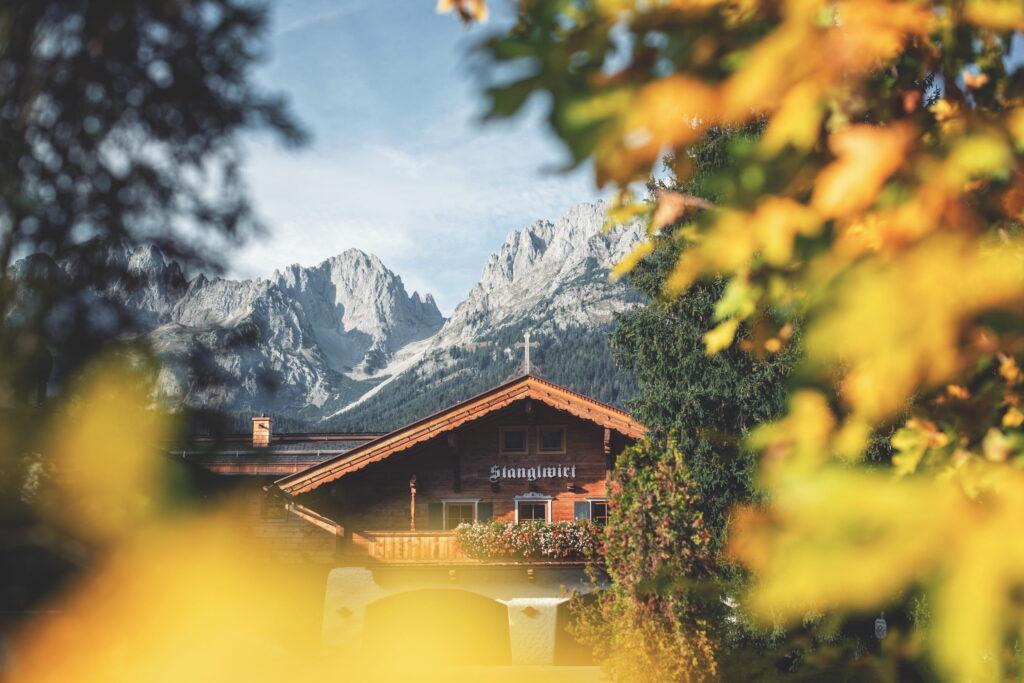 Holzgebäude mit Schriftzug 'Stanglwirt' vor Bergkulisse des Wilden Kaisers im herbstlichen Umfeld mit gelb verfärbtem Laub
