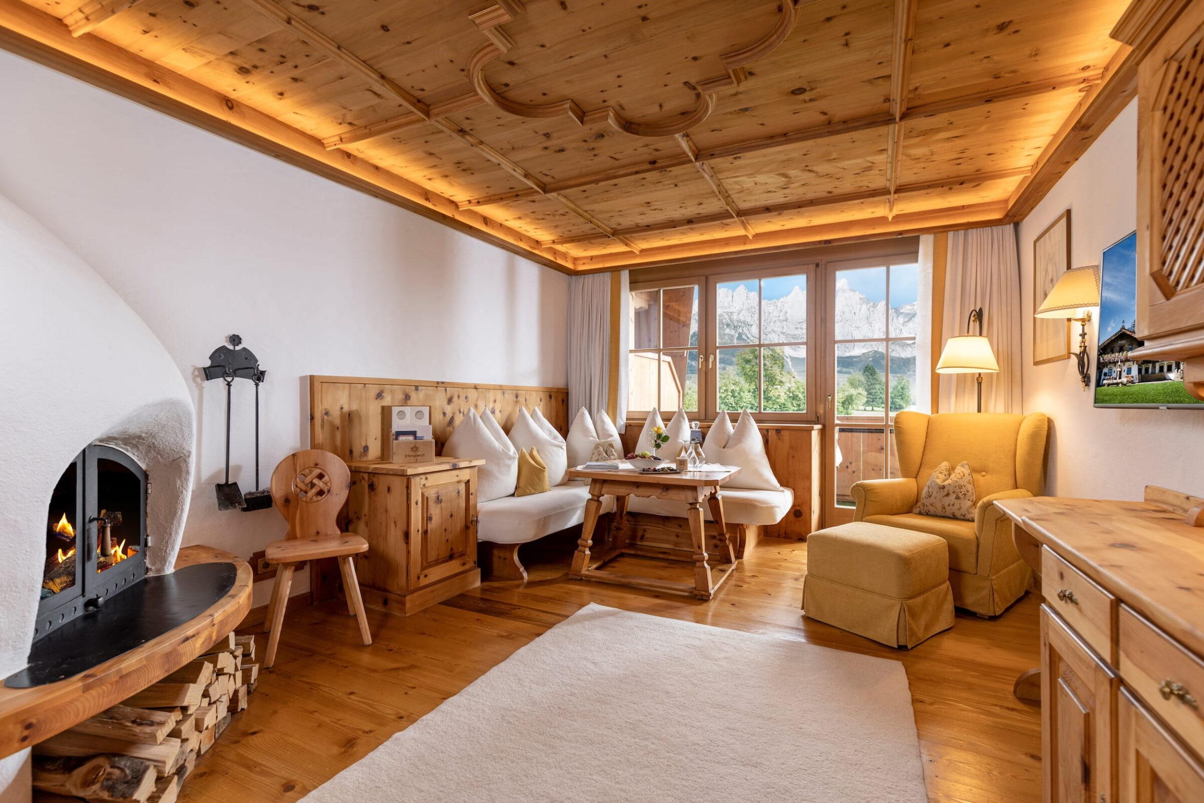 Wohnzimmer der Juniorsuite im Bio- und Wellnessresort Stanglwirt mit Holzboden, Holzdecke, Kamin, Eckbank mit Kissen, gelbem Ohrensessel und großem Fenster mit Blick auf Berge