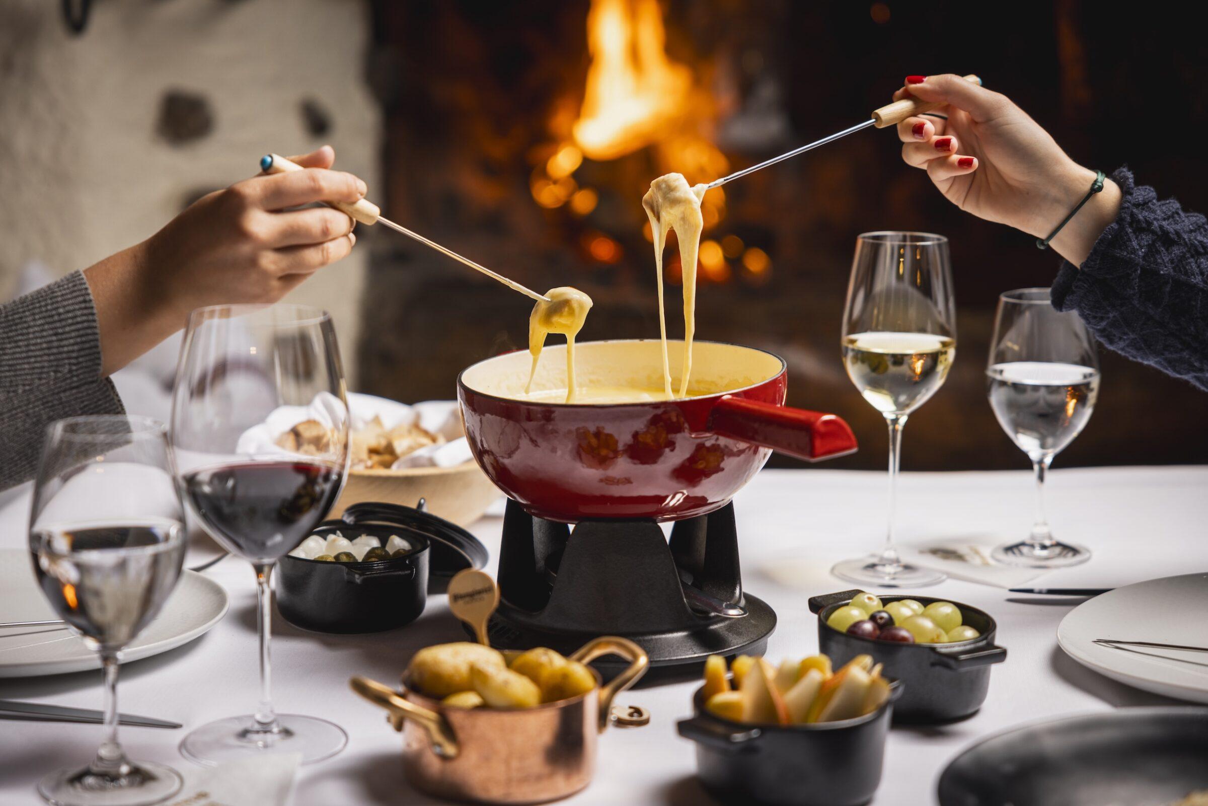 Roter Fonduetopf mit geschmolzenem Käse vor Kaminfeuer im Wellnessresort Stanglwirt in Tirol, zwei Personen tauchen Brotstücke mit Fonduegabeln ein, gedeckter Tisch mit Wein- und Wassergläsern sowie kleinen Schalen mit Beilagen