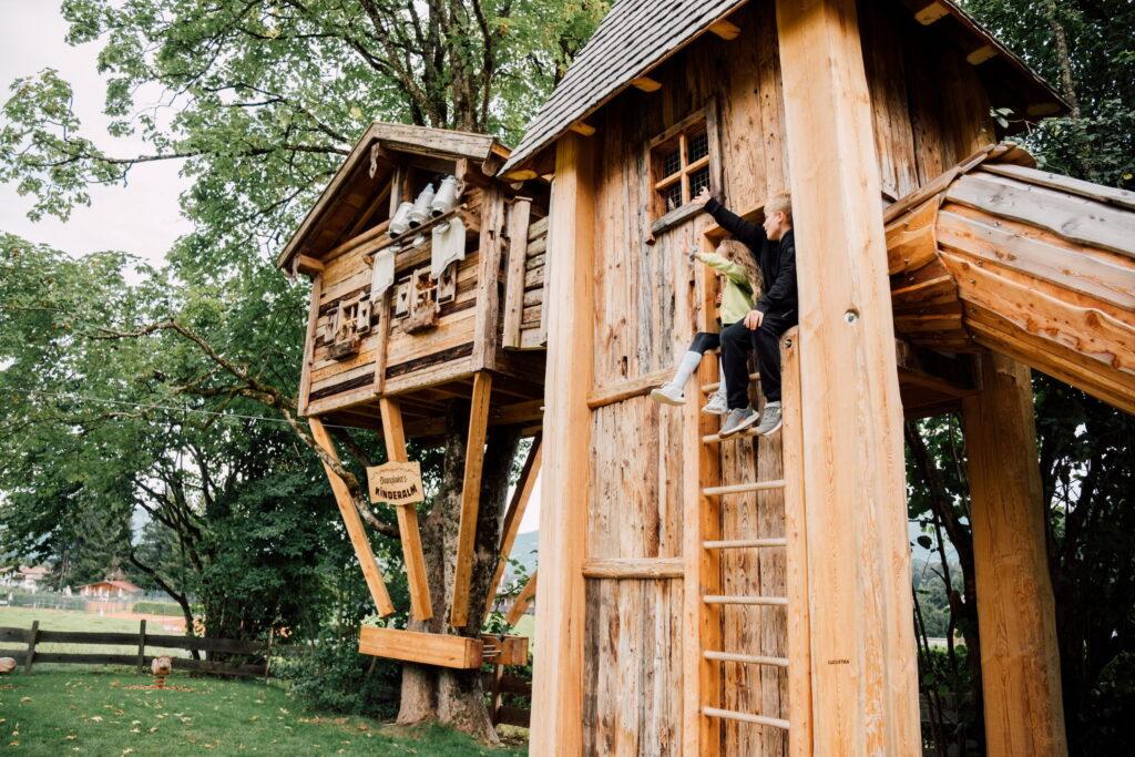 Zwei Kinder sitzen auf einer Holzleiter an einem Baumhaus im Bio- und Wellnessresort Stanglwirt in Tirol