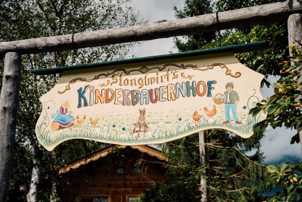 Holzschild mit der Aufschrift 'Stanglwirt's Kinderbauernhof' und einer bemalten Szene mit einem Mann, Hasen und Hühnern, aufgehängt an einem Holzgestell vor Bäumen und einem Holzhaus