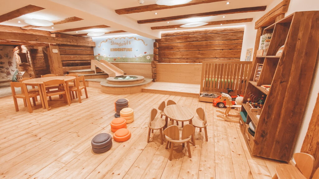 Kinderspielzimmer mit Holztischen, kleinen Stühlen, Sitzkissen, Holzregal mit Spielzeug und Rutsche vor einer Wand mit der Aufschrift 'Stanglwirts Kinderstube' im Bio- und Wellnessresort Stanglwirt in Tirol
