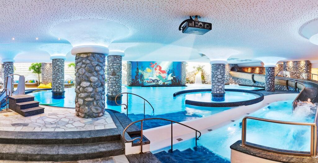 Innenansicht des Kinderwasserwelt-Bereichs im Bio- und Wellnessresort Stanglwirt mit mehreren runden Pools, steinverkleideten Säulen, Wasserrutsche und einer Wand mit einer großen bunten Meerjungfrauenillustration
