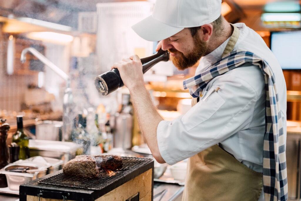 Koch mit weißer Kochmütze und kariertem Geschirrtuch über der Schulter würzt zwei Fleischstücke auf einem Grill im Restaurant des Bio- und Wellnessresorts Stanglwirt in Tirol