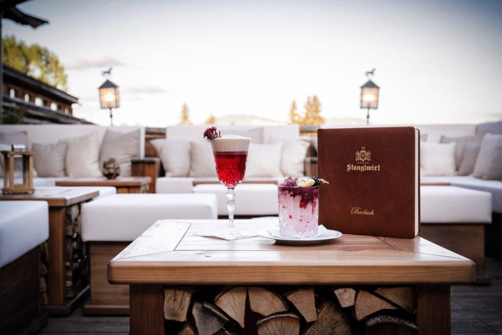 Holztisch mit zwei dekorierten Cocktails und einem braunen Barbuch vor einer Lounge mit weißen Polstern und Kissen im Bio- und Wellnessresort Stanglwirt in Tirol