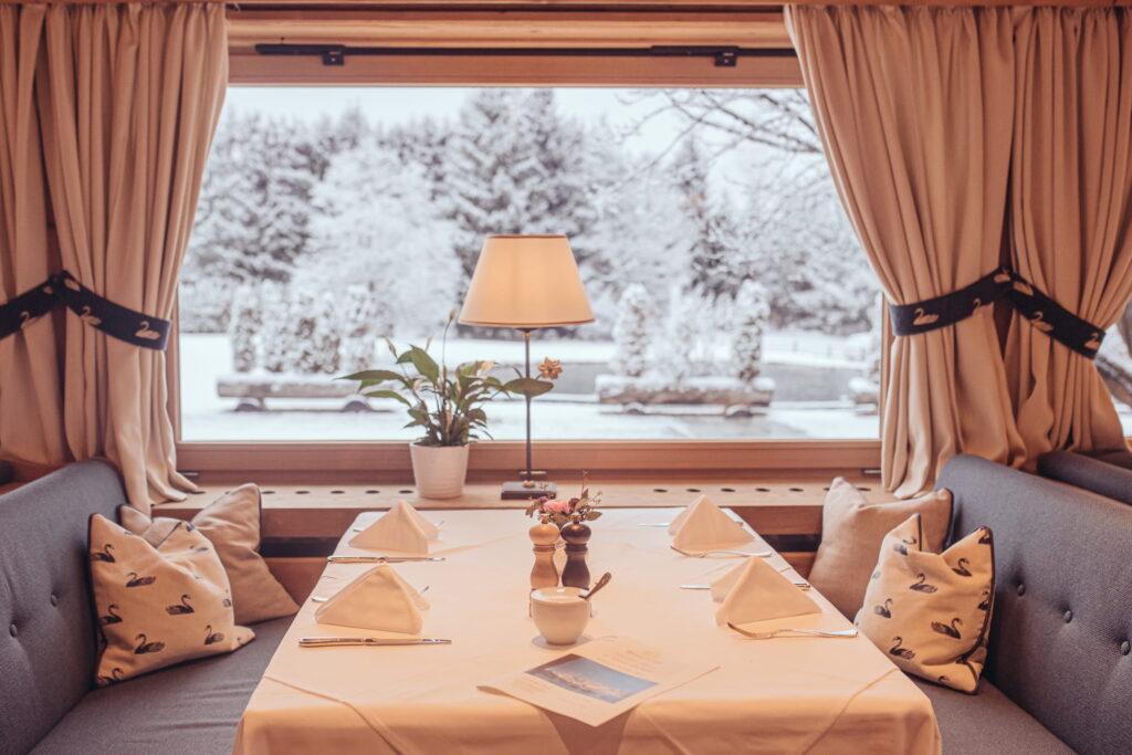 Eingerichteter Esstisch mit weißen Servietten, Kissen mit Schwanenmuster auf Sitzbänken, Tischlampe und Pflanzen vor großem Fenster mit Blick auf verschneite Landschaft Tirols beim Wellnessresort Stanglwirt
