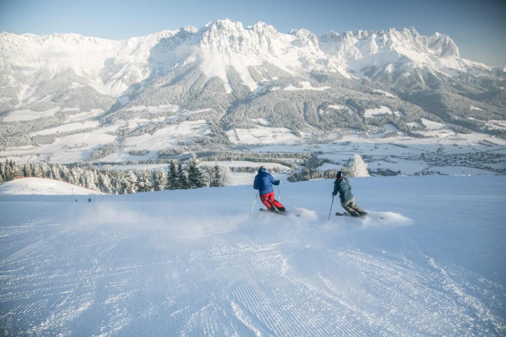 Zwei Skifahrer fahren eine frisch präparierte Piste mit Blick auf schneebedeckte Berge des Wilden Kaisers in Tirol