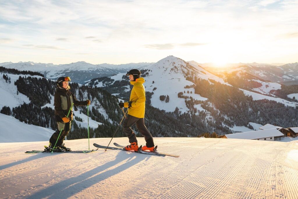 Zwei Skifahrer in Winterkleidung stehen auf einer frisch präparierten Skipiste mit Blick auf schneebedeckte Berge der Kitzbüheler Alpen und Sonnenuntergang im Hintergrund