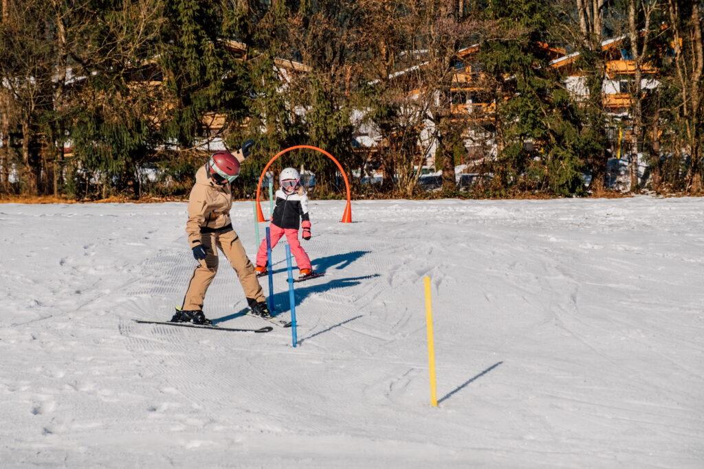 Zwei Personen in Skikleidung, eine Erwachsene und ein Kind, üben Skifahren auf einer Schneefläche mit Trainingsstangen und einem orangen Tor vor einem Wald