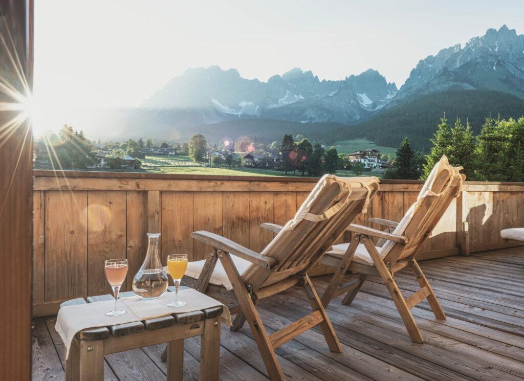 Eine sonnenüberflutete Holzterrasse mit zwei Liegestühlen und Blick auf majestätische Berge der Kitzbüheler Alpen in Tirol. Auf dem Tisch stehen Getränke bereit, was eine einladende Atmosphäre schafft. Bio- und Wellnessresort Stanglwirt