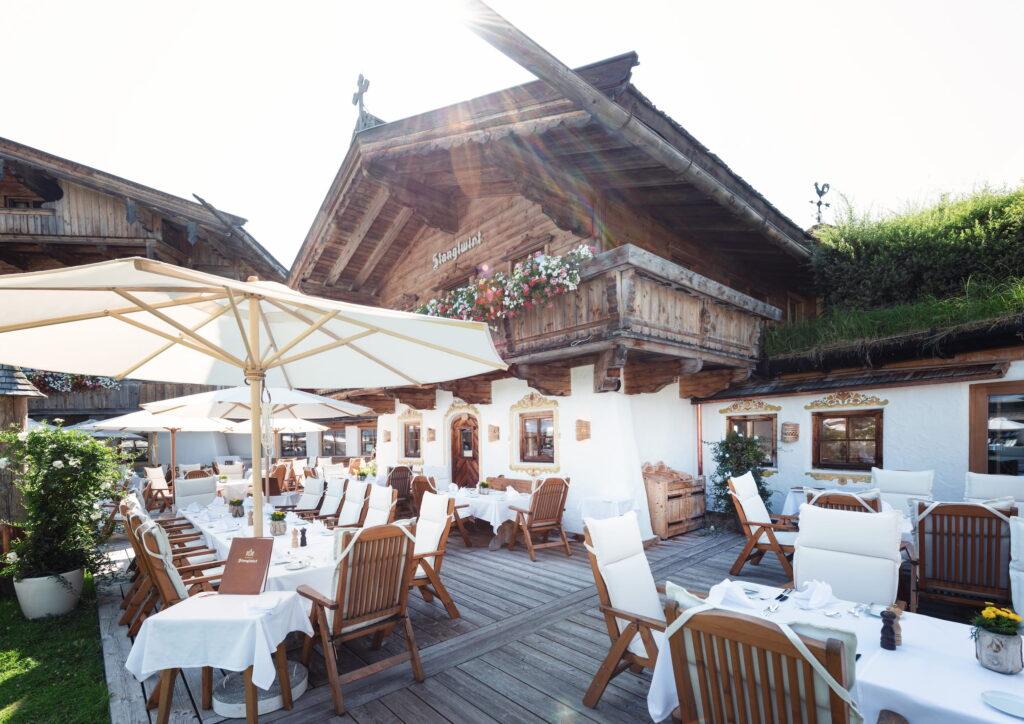 Terrasse mit Holztischen und -stühlen mit weißen Polstern und Sonnenschirmen vor einem traditionellen Tiroler Holzhaus mit Balkon und Blumen am Bio- und Wellnessresort Stanglwirt