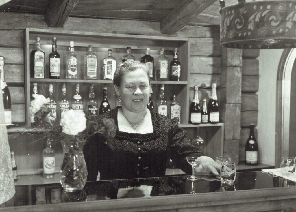 Frau in traditioneller Tracht steht hinter einer Bar mit Regalen voller Spirituosenflaschen und einem Glas mit Aufschrift 'Stanglwirt'