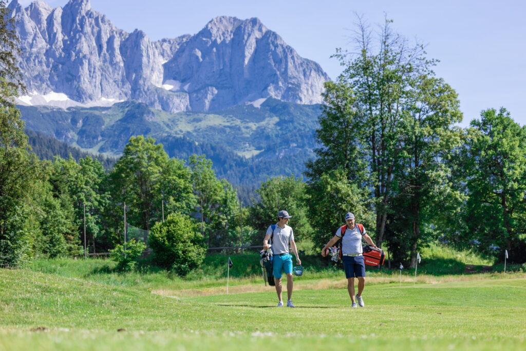 Zwei Männer mit Golfbags gehen auf einem Golfplatz vor bewaldeten Bergen und Felsgipfeln des Wilden Kaisers im Hintergrund