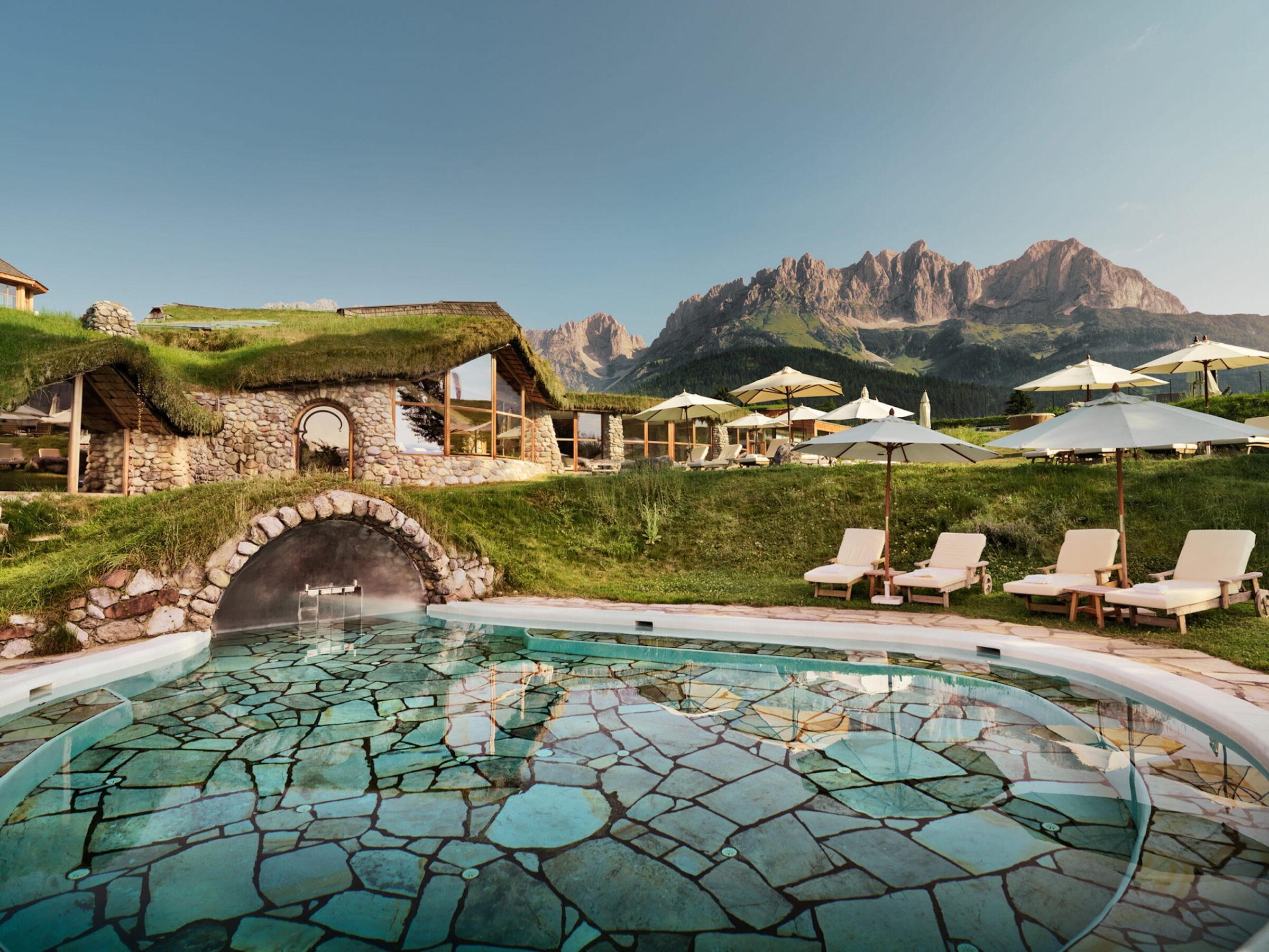 Außenpool im Bioresort Stanglwirt in Tirol mit Natursteinboden und Steinbogen, umgeben von Liegestühlen mit Sonnenschirmen vor grasbewachsenen Gebäuden und Bergkulisse des Wilden Kaisers