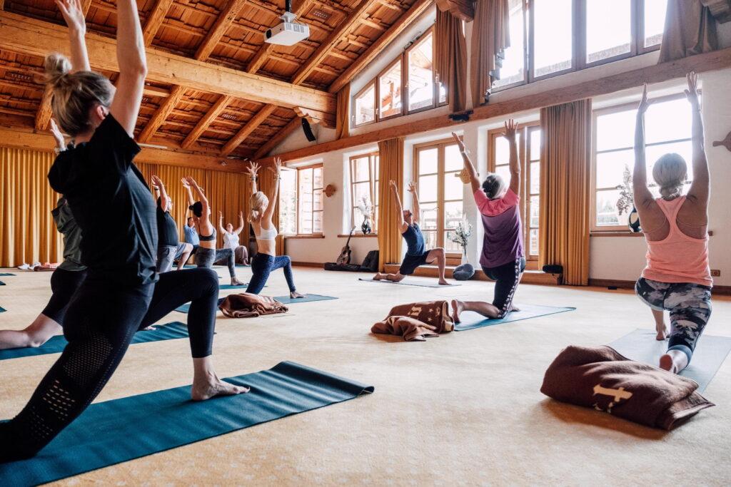 Ein Yoga-Kurs in einem geräumigen, sonnendurchfluteten Studio mit Holzbalken im Bio- und Wellnessresort Stanglwirt am Wilden Kaiser in Österreich. Die Teilnehmer strecken sich auf ihren Matten nach oben und schaffen so eine ruhige und konzentrierte Atmosphäre.