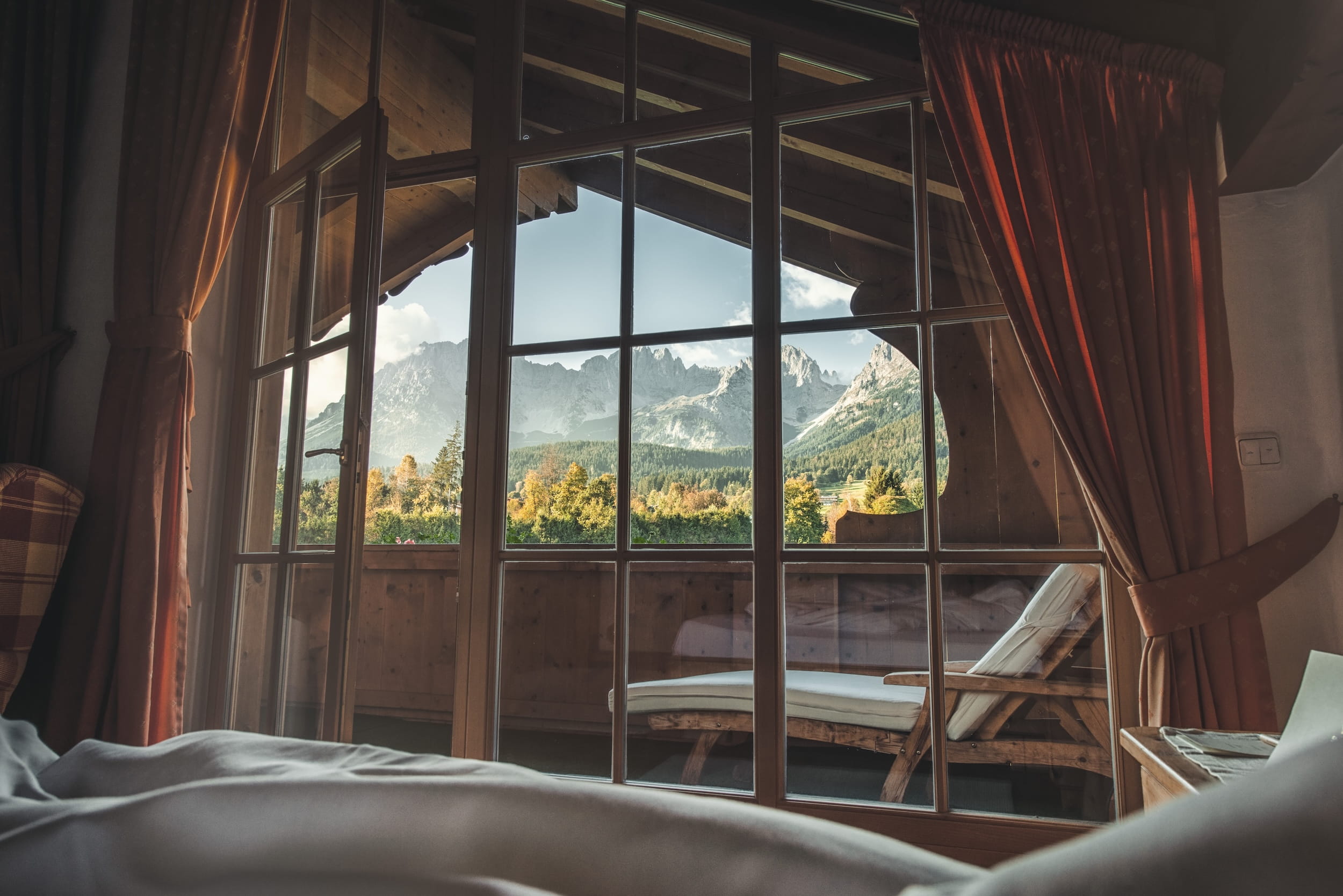 Blick aus einem Zimmer des Bio- und Wellnessresorts Stanglwirt in Tirol durch eine verglaste Balkontür auf eine Liege und die Berge des Wilden Kaisers