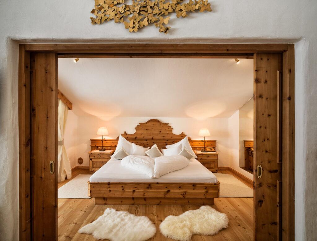 Ein gemütliches Schlafzimmer im Bio- und Wellnessresort Stanglwirt mit einem rustikalen Holzbett und Nachttischen, ausgestattet mit weißer Bettwäsche. Sanfte Beleuchtung, Fellteppiche und Holztüren sorgen für eine warme, einladende Atmosphäre.