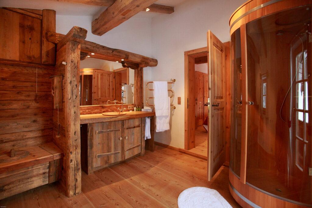 Rustikales Badezimmer aus Holz im Chalet Hüttling Moos des Bioresorts Stanglwirt in Tirol mit sichtbaren Balken und Sauna. Warme Farbtöne, ein Waschtisch im Blockhausstil, eine offene Tür, Handtuchhalter und eine gemütliche Atmosphäre.