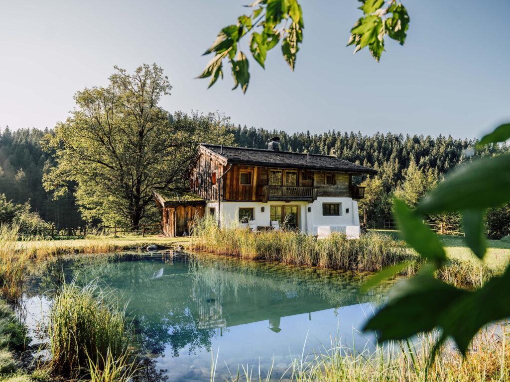 Das rustikale Chalet Hüttling Moos des Bio- und Wellnessresorts Stanglwirt in Tirol an einem ruhigen Teich, umgeben von üppigem Grün und mit einem Wald als Kulisse. Das Sonnenlicht fällt durch die Bäume und schafft eine friedliche Atmosphäre.
