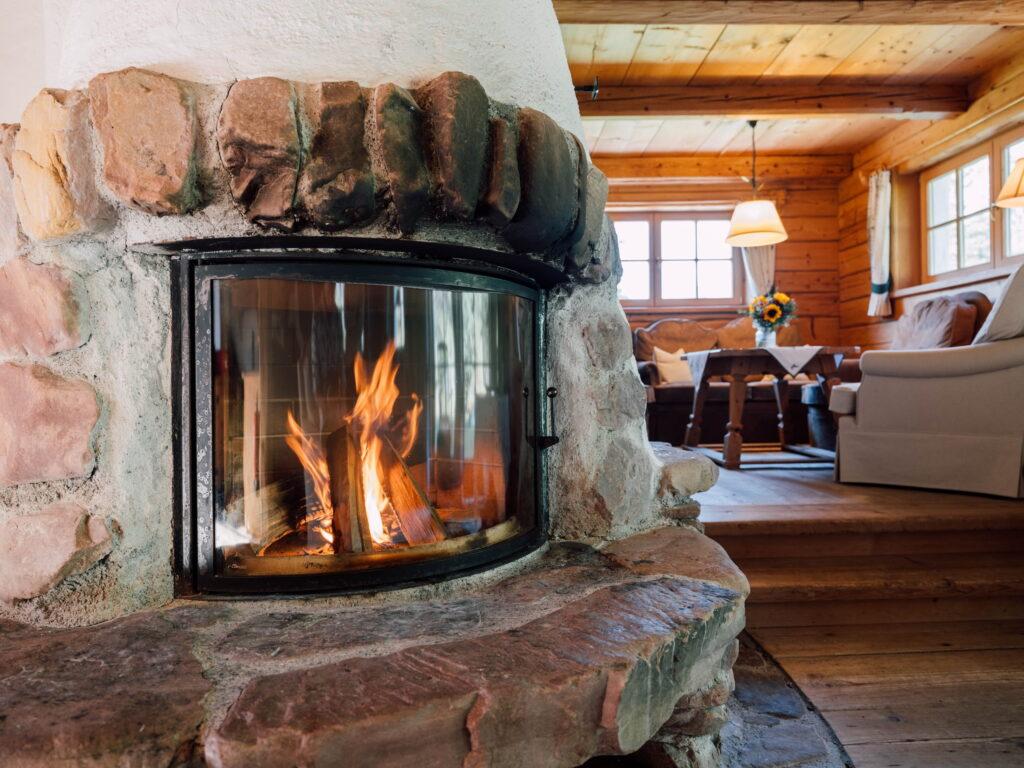Ein gemütliches Wohnzimmer im Chalet Hüttling Moos des Stanglwirts am Wilden Kaiser mit einem warm leuchtenden Steinkamin, Holzwänden, weichen Sofas und einem Tisch mit Sonnenblumen, das eine rustikale, einladende Atmosphäre schafft.