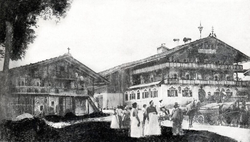 Historische schwarz-weiße Aufnahme des Gasthauses Stanglwirt mit mehreren Personen in traditioneller Kleidung und Pferdekutschen davor