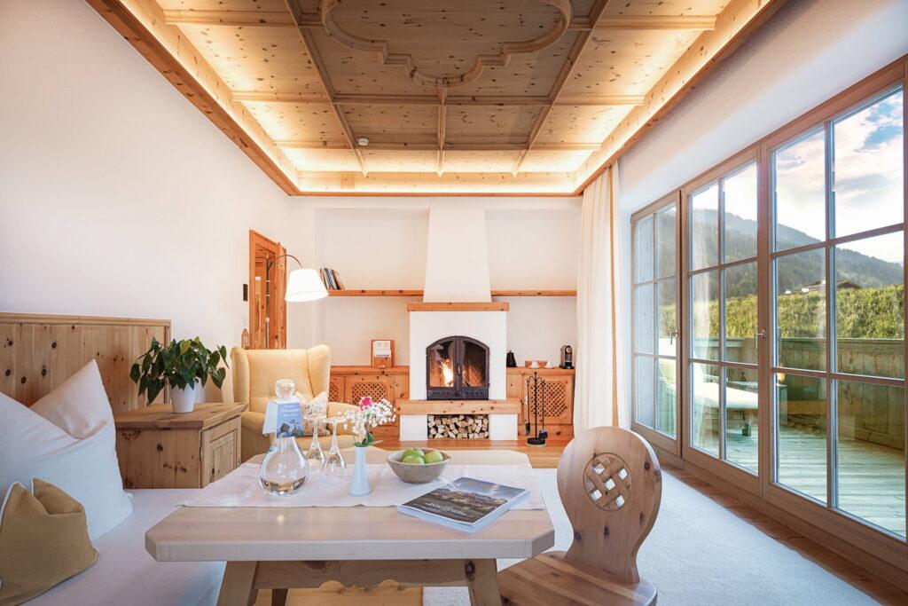 Gemütliches Wohnzimmer mit Holzdecke und Holzmöbeln sowie einem befeuerten Kamin. Durch die großen Fenster bietet sich ein malerischer Blick auf die Kitzbüheler Alpen, der eine warme, einladende Atmosphäre schafft. Im Bio- und Wellnessresort Stanglwirt in Österreich.